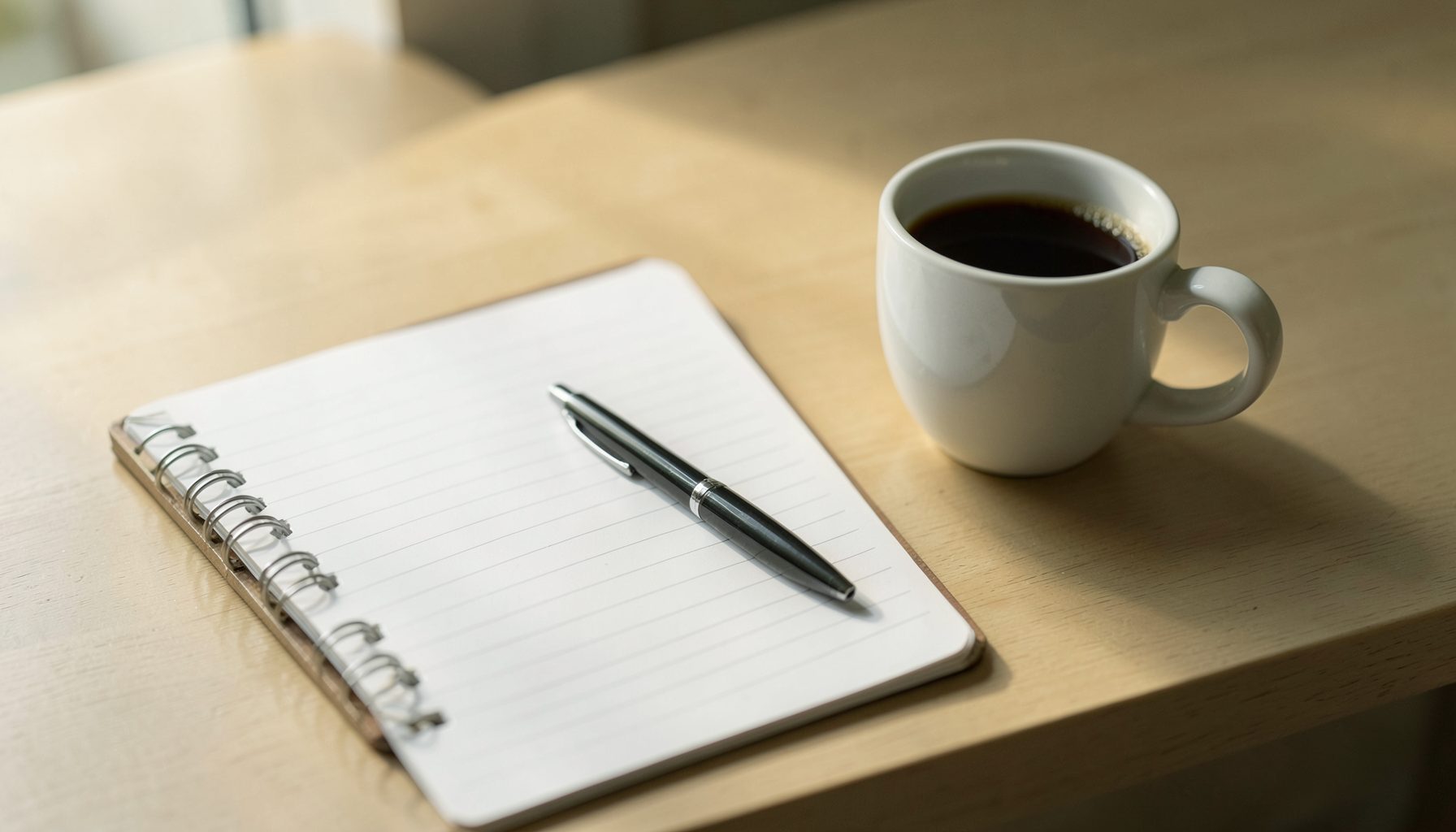 A clean desk with a notebook, pen, and a cup of coffee in soft morning light, warm beige and muted green tones